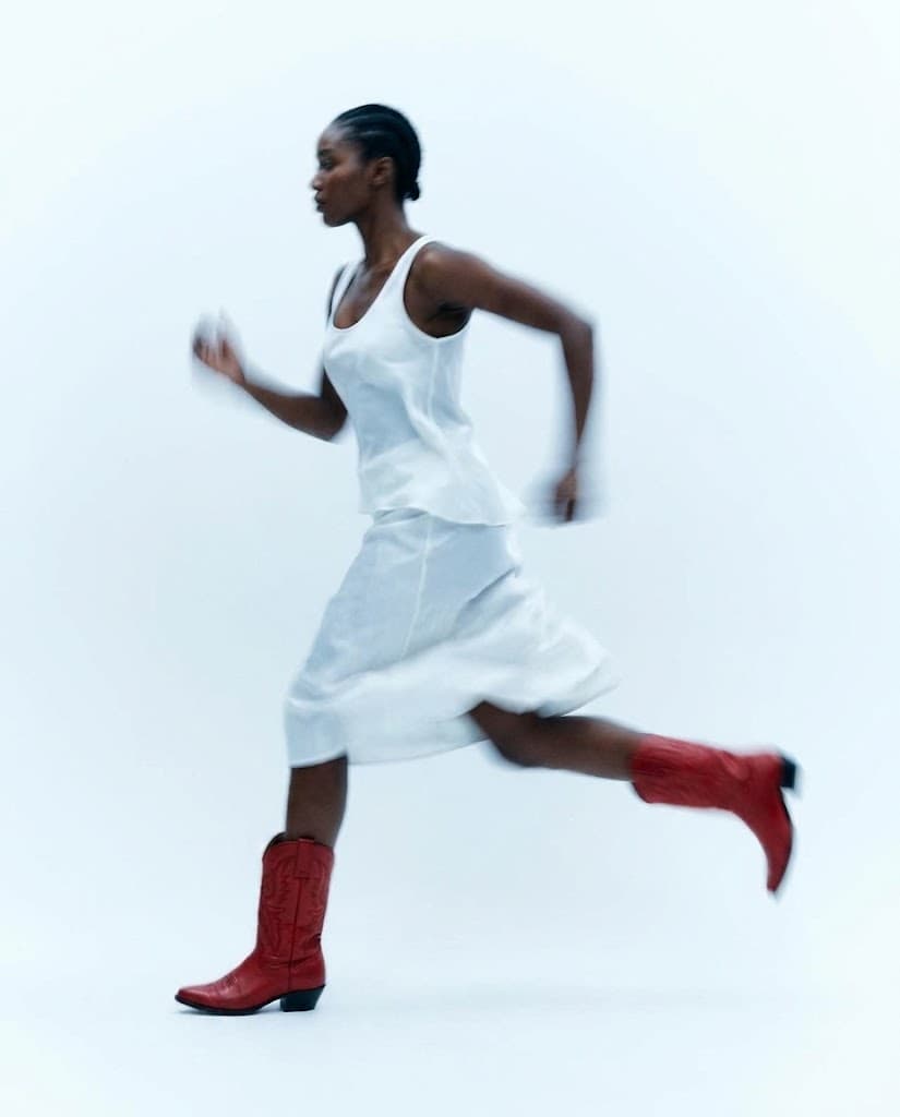 A Black woman running in a white dress and red cowboy boots, moving through cool daylight.